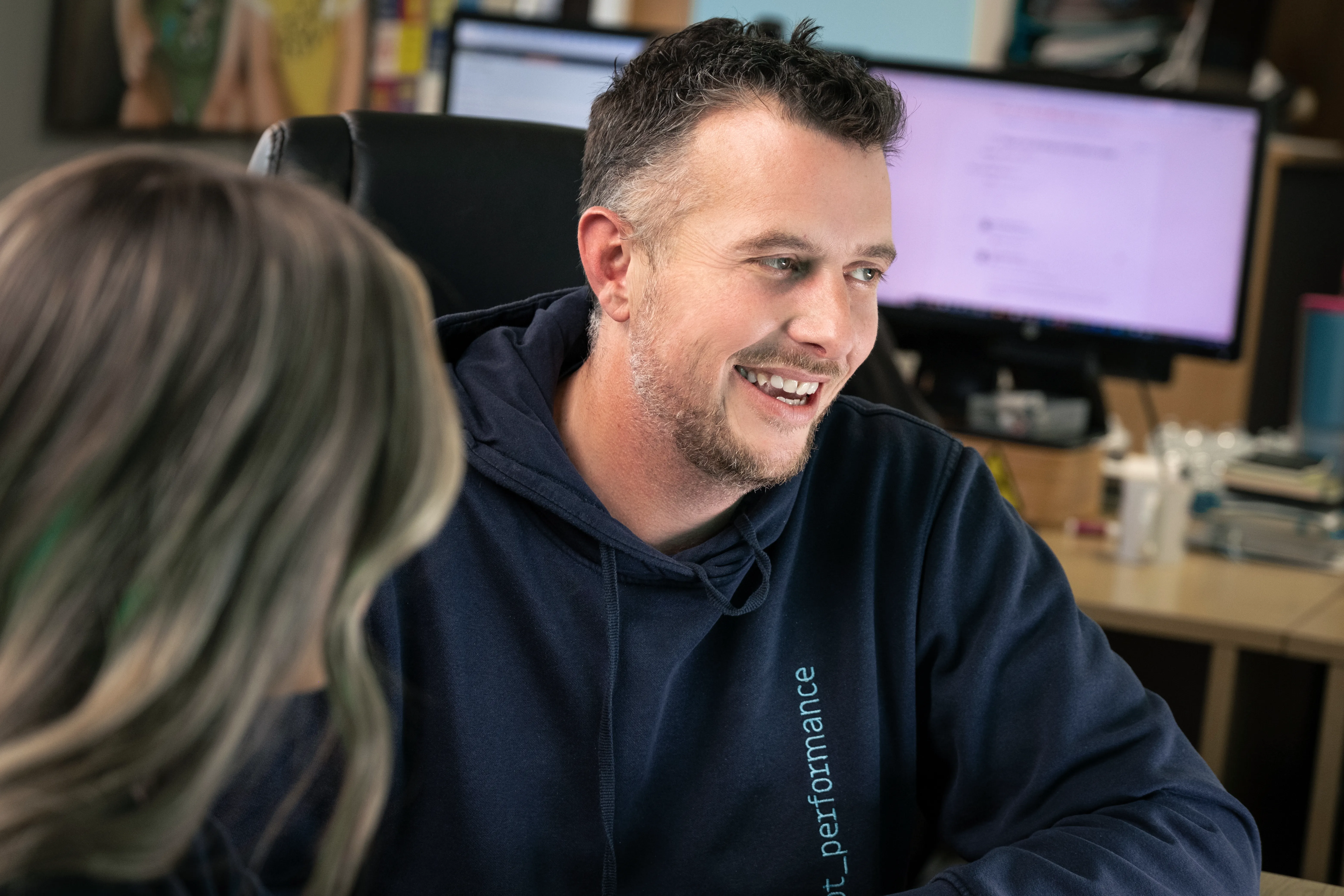 A smiling man in a dot_performance hoodie listens during a conversation with a colleague in a modern office environment with screens visible in the background.